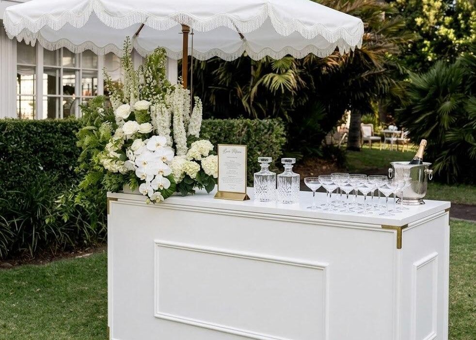 Bar extérieur blanc élégant avec parasol à franges, compositions florales et verres à cocktail, pour réception de mariage en plein air.