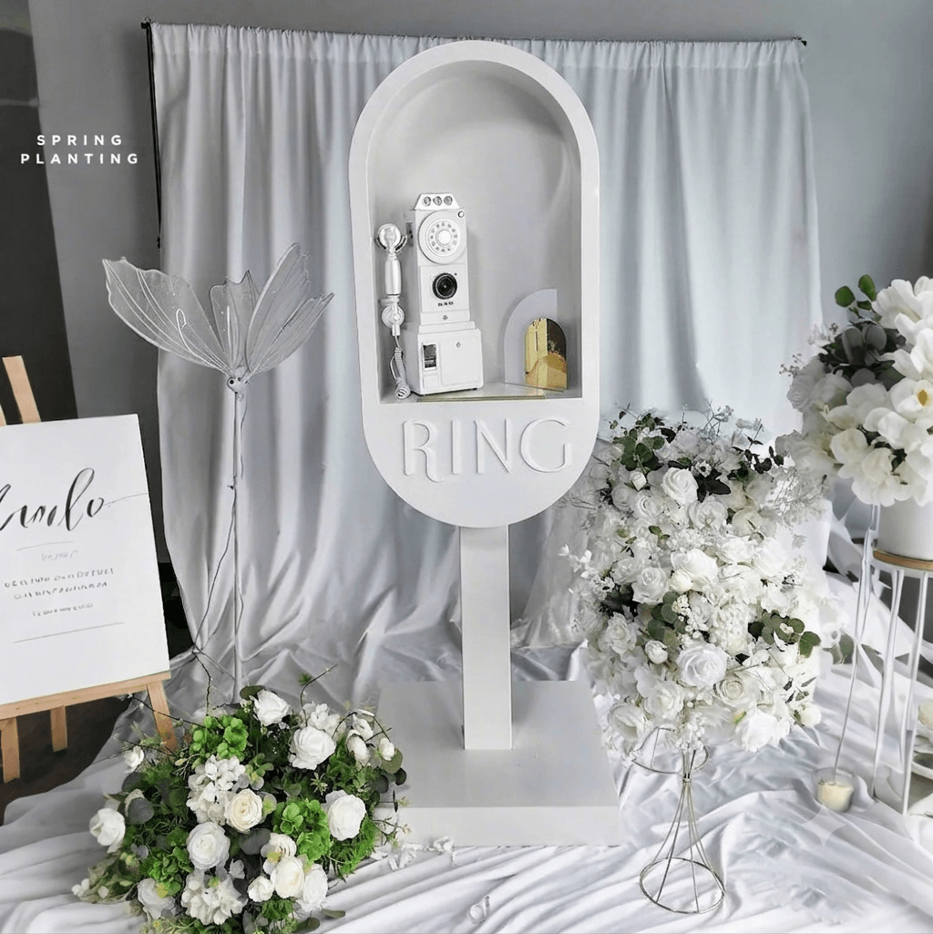 Cabine téléphonique vintage blanche avec téléphone à cadran, entourée de roses blanches, utilisée comme livre d'or audio pour un mariage.